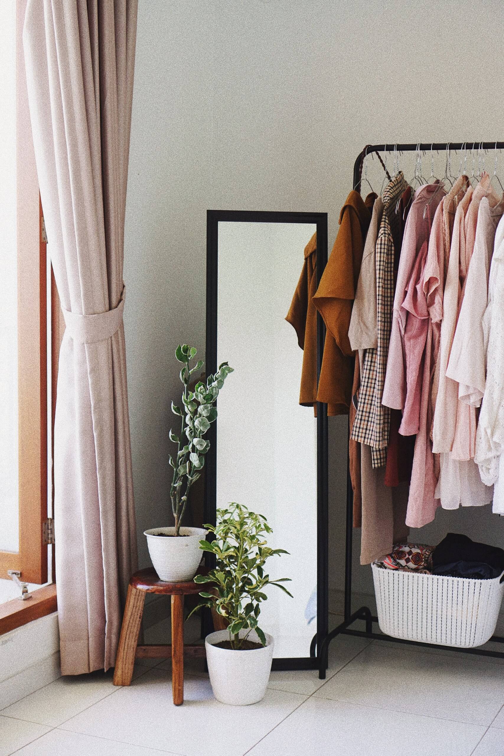 detalhe de quarto feminino com cabide com roupa em tons de cor-de-rosa, espelho, plantas e cortinado rosa 