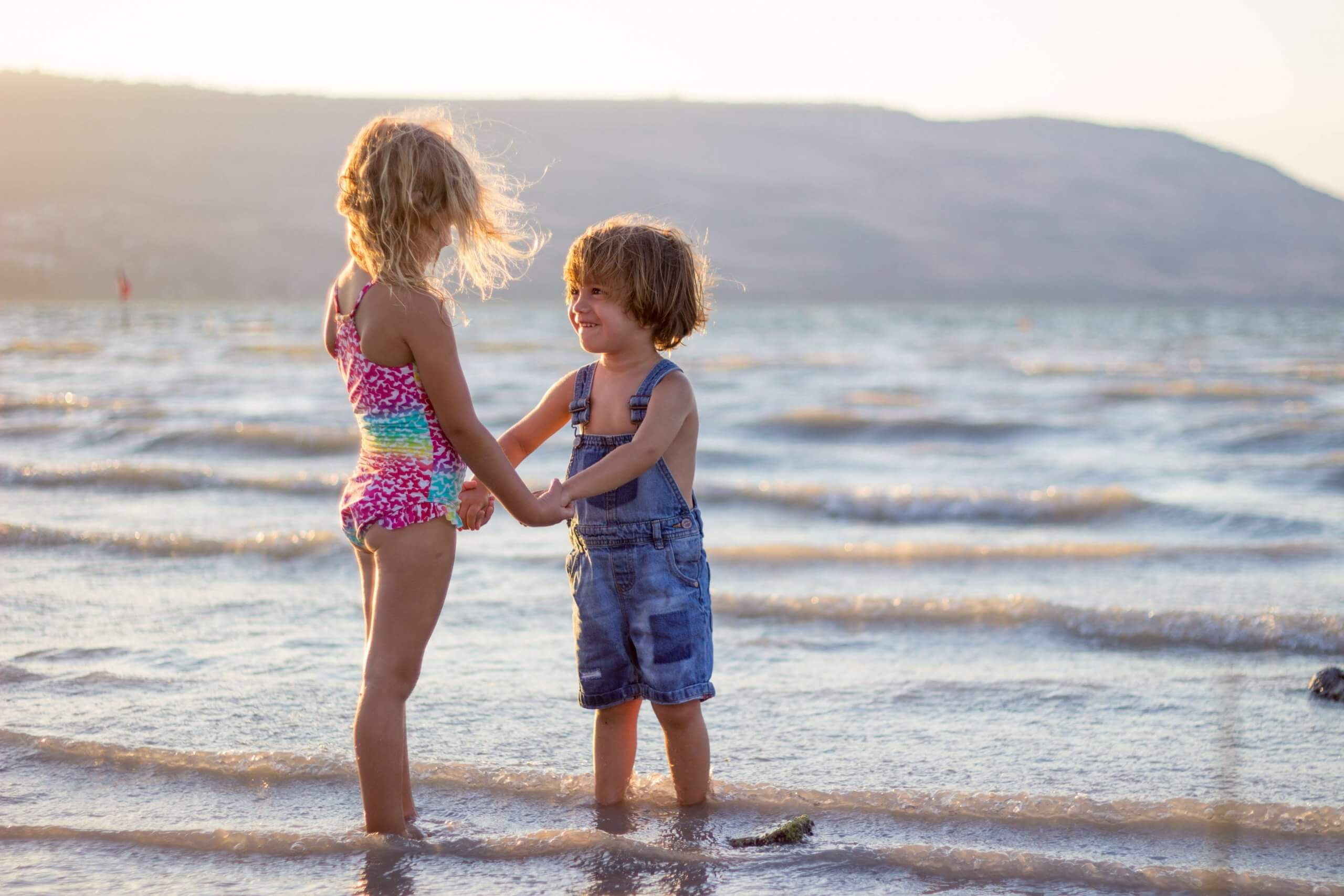 menina e menino de mãos dadas a olhar um para o outro na praia com os pés no mar